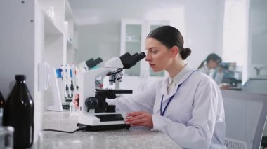 Genç çekici bir kadın masada oturuyor ve mikroskoptan bakıyor. İçeride çalışan güzel görünümlü kadın laboratuvar çalışanı görüntüsü. Gelişmiş laboratuvarda biyoteknoloji araştırması. Aşı