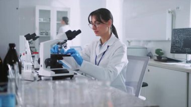 Modern laboratuarda çalışırken, eldiven giyen, ekipman kullanan ve mikroskop altında bakan güzel bir genç kadın görüntüsü. Beyaz laboratuvar önlüklü beyaz kızın portresi iş yerinde oturuyor.