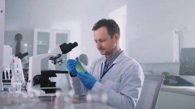 Modern mikroskobun önünde oturan ve laboratuvarda üzerinde yeşil madde bulunan laboratuvar kavanozu tutan beyaz laboratuvar önlüğü giyen ciddi odaklanmış genç bir beyaz adam. İş günü. Araştırma konsepti
