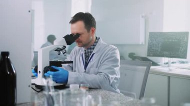 Beyaz önlüklü genç laboratuvar teknisyeni örneklere bakıyor ve laboratuvarda mikroskop kullanıyor. Mavi eldivenli adam iş başında. Biyomedikal araştırma. Bilim konsepti. Kapalı