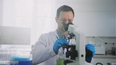 Beyaz önlüklü erkek bilim adamının laboratuvarda çalışırken mikroskoba baktığı görüntüler. İşyerinde mavi eldivenli, odaklanmış genç bir adam. Modern bir laboratuarda. Örnek çalışma. Kapalı