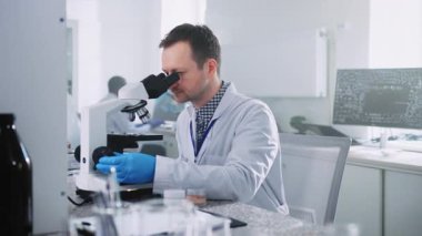 Laboratuvarda deney sırasında mikroskoptan bakan tıraşsız beyaz bir adamın portresi. Beyaz işçi kıyafetli bir erkek bilim adamı aşıyı test ediyor. İçeride. Araştırmacı işi konsepti. Mikrobiyoloji