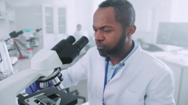 Afrika kökenli Amerikalı üniformalı bir bilim adamı mikroskop altında araştırma yapıyor. Teknolojik ekipmanlarla modern bir laboratuvarda çalışan bir mikrobiyolog.