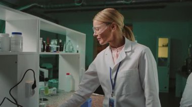 Beyaz önlüklü, koruyucu gözlüklü, beyaz beyaz tenli bir kadının elinde test tüpü tutarken iş arkadaşının akşamları modern tıp araştırma laboratuvarında notlar aldığı görüntüler. Bilimsel