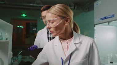Bilim adamı ve asistanının akşamları laboratuvarda çalıştığı görüntüler. Bir kadın deney tüpünü tutuyor ve erkeğe veriyor. Odaklanmış adam not alıyor. Kapalı