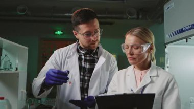 Bilim adamları deney sonuçlarını tartışıyor. Doktor ve asistan konuşuyor. Beyaz önlüklü bir adam deney tüpü tutuyor ve kadın gece modern laboratuarda notlar alıyor. Takım çalışması. Araştırma.