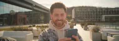 Akıllı telefon kullanan, dışarıda sohbet eden mutlu ve çekici genç bir adamın portresi. Sakallı adam güzel modern eğlence merkezine bakıyor. Açık havada. Anamorfik mercekle çekilmiş.