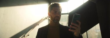 İş kıyafetleri içinde, elinde akıllı telefon tutan ve modern ofis binasında sohbet eden kendinden emin beyaz bir adamın portresi. İş adamı bir aletle mesaj atıyor. Güneş ışığı. Anamorfik lens patlaması. Kapalı
