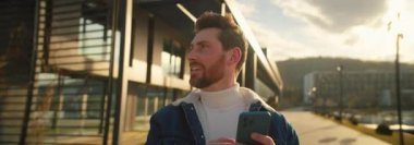 Genç, sakallı, akıllı telefon kullanan ve güneşli bir günde lüks seyahat merkezinde tatilin tadını çıkaran yakışıklı bir adam. Yaşam tarzı konsepti. Dağlarda gölü olan modern bir otel. Anamorfik çekim