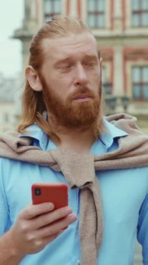 Bakımlı sakallı, uzun saçlı şık bir adam akıllı telefonunu açık havada kontrol ediyor.