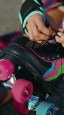 Şık bir kadın paten yapmaya hazırlanıyor, patenlerini bağlıyor, şapka ve eldiven giyiyor, elinde bir kaykay tutuyor ve neşeyle gülümsüyor.