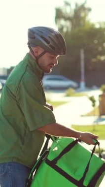 Yeşil kıyafetli ve kasklı bir bisikletçi kalabalık bir şehir bölgesinde dolaşırken bir sırt çantası taşıyor.