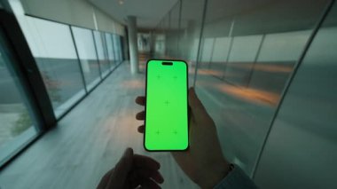 El adamı yeşil ekranlı dikey telefonu tutuyor ve kaydırıyor. Arka planda büyük pencereli modern alan. Akıllı telefondan yazıyorum.