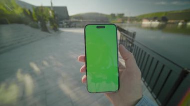 Yeşil ekranlı, el ele tutuşan ve dikey olarak telefonu kaydıran adama bakın. Arka plandaki gölde, güneş ışığında. Açık havada akıllı telefondan yazıyorsun.