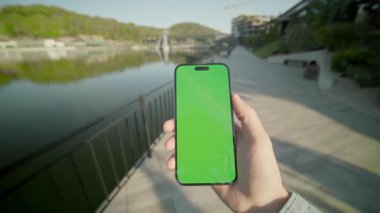Açık havada çalışan adam yeşil ekran yürüyüşlü, dikey telefonu tutuyor ve kaydırıyor. Arka plandaki gölde, güneş ışığında. Akıllı telefondan yazıyorum.