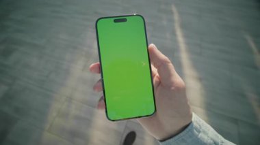 El ele tutuşan ve yeşil ekranlı dikey telefonları kaydıran adamı kapat. Arka planda güneş ışığı. Akıllı telefondan yazıyorum.