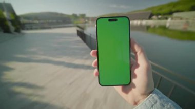 Eller dikey olarak yeşil ekranda yürüyen akıllı telefon cihazını tutuyor. Arka plandaki gölde, güneş ışığında. Doğa