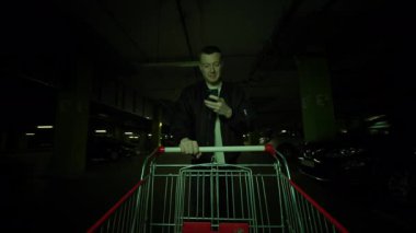 Alışveriş arabalarıyla yürüyen çekici genç adam geceleri alışveriş merkezinin yeraltı otoparkında gülümseyen bir telefon kullanıyor.