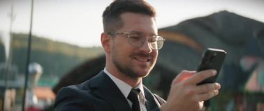 Siyah takım elbiseli, gülümseyen sakallı, akıllı telefon tutan ve sohbet eden genç bir adamın yatay görüntüsü. Parkın bulanık arka planında gözlüklü beyaz iş adamının portresi. Açık havada. Bahar