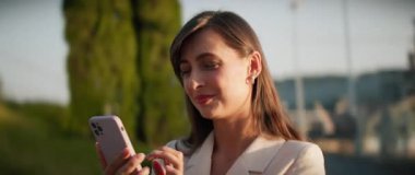 Güzel görünümlü, kahverengi saçlı, elinde akıllı telefon tutan ve sohbet eden genç bir kadın. Çekici beyaz bir kızın öğle arasında telefonda mesajlaşması ve gülümsemesi. Güneşli bir gün. Açık havada.