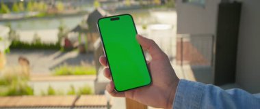 Genç adam elinde telefonla pencerenin yanında dikey yeşil ekran duruyor. Arka sokakta.