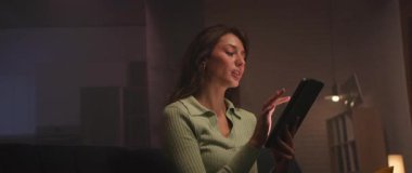 Kanepede oturan çekici beyaz bir kadının yatay görüntüsü. Gayri resmi kıyafetli çekici kız oturma odasında dijital tablet tutuyor. Akşam vakti. Kapalı.