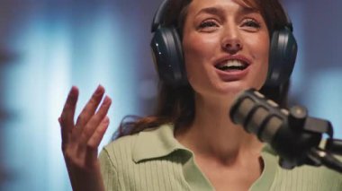 Stüdyoda mikrofonlu ve kulaklık takıp podcast kaydeden genç bir bayan radyo sunucusu. Güzel görünümlü, gülümseyen bir kızın görüntüsü. Online radyo konsepti. Akşam vakti. Dijital içerik