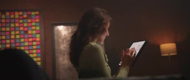 360 derece hızlandırılmış ev. Dijital tablet taşıyan, sosyal medyada gezinen, mesajlar okuyan, evde sohbet eden beyaz bir kadın. Güler yüzlü kız akşamları alet kullanıyor.