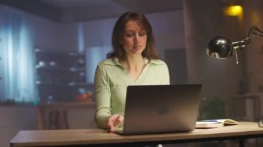 Laptopun önünde oturup klavyede daktilo çalan, günlük kıyafetlerle odaklanmış beyaz bir kadının görüntüleri. Akşamları başarılı bir kız çalışıyor. Güzel, modern iç mekan. İz sürme