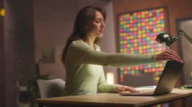 Çekici odaklı genç bir kadın masa başında oturuyor ve akşam çalışmak için dizüstü bilgisayar açıyor. Evden çalışan güzel görünümlü beyaz bir kızın portresi. Samimi bir daire. Çekim sırasında iz sürüyorum. Uzak