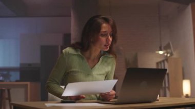 Laptop başında oturan ve oturma odasında diyagramlarla çalışan odaklanmış, yakışıklı, beyaz bir kadının 360 derecelik hızlandırılmış görüntüsü. Akşam vakti. Modern daire. Modern iç mekan. İz sürme