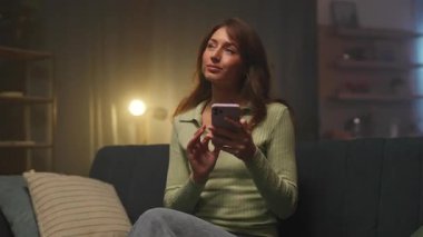 Oturma odasındaki kanepede otururken akıllı telefon kullanan günlük kıyafetlerle çekici, neşeli genç bir kadın. Güzel görünümlü mutlu beyaz kız projesini kutluyor. Gece vakti. İz sürme