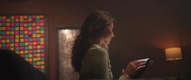 Akşamları oturma odasındaki kanepede otururken siyah tablet kullanan güzel kahverengi saçlı genç bir kadının görüntüsü. Odaklanmış güzel bir kızın ekrandaki cihaza dokunuşu. Kamera hareket ediyor