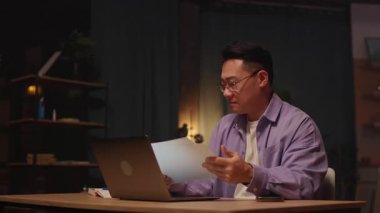 Gözlüklü Asyalı iş adamı dizüstü bilgisayar kullanıyor ve telefonda konuşuyor. Geceleri ekrana ciddi ciddi bak. Kağıt üzerinde çalışmak. 360 derece. İz sürme görüntüsü etrafında hareket