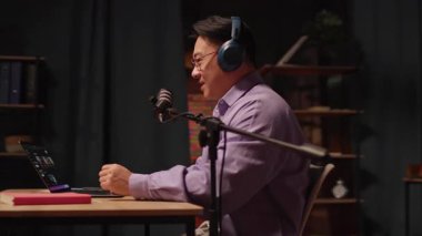 Asyalı içerikli yaratıcıyı kapat, dizüstü bilgisayarda konuşan kulaklıkla ve gece yayın stüdyosunda mikrofon röportajı yapan adam. - Eve. 360 derece. Kamera hareket ediyor