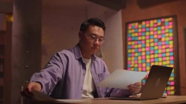 Gözlüklü Asyalı işadamı bilgisayarla kağıt kullanıyor. Geceleri ekrana ciddi ciddi bak.