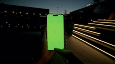 Erkek ellerinin akıllı telefonu tutarken ve dışarıdaki yeşil ekrana dokunurken yakın çekim görüntüleri. Genç bir adam gecenin karanlığında merdivenlerin arka planında el kol hareketi yapıyor. Krom anahtar. Mobil teknoloji