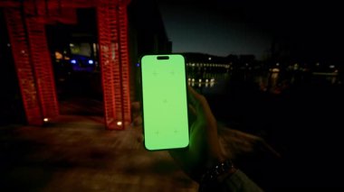 Erkek, akıllı telefonu dikey modda tutuyor. Geceleri modern tatil köyünün bulanık arka planında yeşil ekranı taklit ediyor. Karanlıkta cep telefonu kullanan tanımlanamayan beyaz adam. Işıklandırma