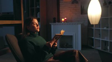 Akşamları sandalyeye oturmuş kitap okuyan genç ve güzel bir kadının manzarası. Romantizme odaklanmış. Evde, şöminenin yanında dinlen.