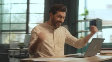 Masada oturan mutlu iş adamı ekranda dizüstü bilgisayar kullanıyor gülümsüyor ve ofiste dans ediyor. Tatmin oldum. Kamera hareket ediyor. 360 Derece