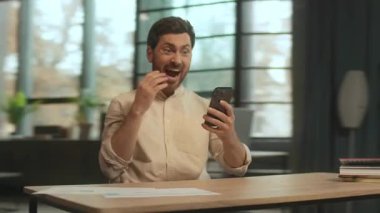 Masada oturan mutlu sakallı bir adam sosyal medyada yazarken gülümsüyor ve şarkı söylüyor. Modern ofis. Kamera hareket ediyor. 360 Derece