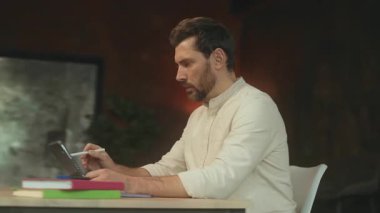 Masada oturan yaratıcı sakallı adam ofisinde kağıtlarla tablet bilgisayar kullanıyor. Düşünüyorum. Kamera hareket ediyor. 360 Derece