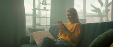 Evdeki kanepede oturan mutlu genç kadın telefon kullanarak gülümsemeyi hisseder. Odada güneş ışığı var. Kamera hareket ediyor. 360 derece