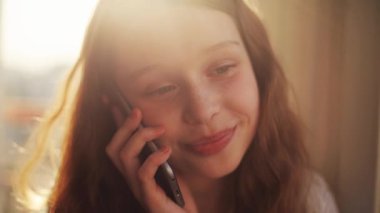Genç bir kız, mutlu bir şekilde akıllı telefonuyla sohbet ediyor. Yüzü, sıcak ve davetkar doğal ışıkla aydınlanıyor.