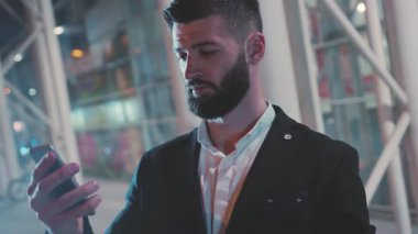Şık bir adam telefonunu modern bir şehir ortamında kontrol eder. Teknoloji ve gece hayatını ön plana çıkarır..