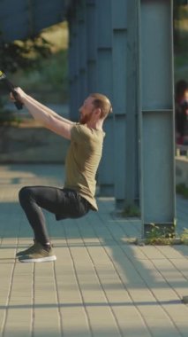 Bir adam açık hava spor salonunda çömeliyor, gücünü ve çevikliğini bir sporcu gibi sergiliyor.