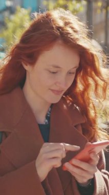 Bu videoda, kızıl saçlı neşeli bir kadın cep telefonuyla meşgul oluyor, neşeli ifadeler sergiliyor ve dijital etkileşimlerden zevk alıyor.