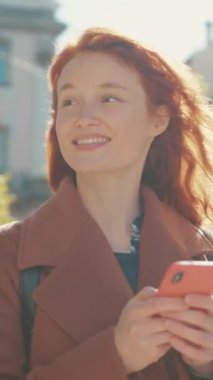 Canlı kızıl saçlı neşeli bir kadın cep telefonunu kullanıyor. Duygularını meraktan zevke yansıtıyor. Modern iletişimi vurguluyor.