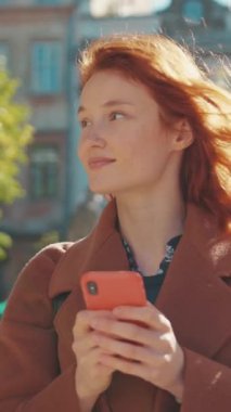 Neşeli kızıl saçlı bir kadın cep telefonuyla meşgul oluyor, mesajları kontrol ederken gülümsüyor, modern sosyal etkileşimdeki neşe ve bağlantıyı yansıtıyor.