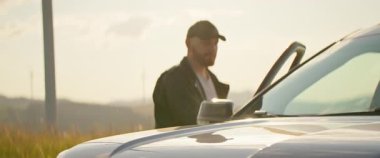 Şapkalı, siyah deri ceketli, kızıl sakallı genç bir adamın araba kapısını kapatıp etrafa bakınışının yatay görüntüsü. Güneşli bir gün. Alan